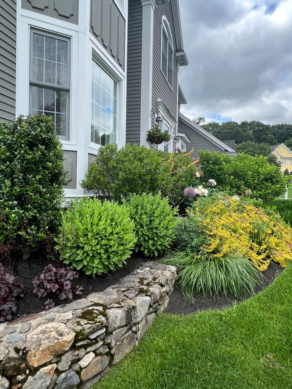 Gray house with large windows, surrounded by lush green shrubs and colorful flowers. Stone wall and grassy lawn in foreground, cloudy sky above.
