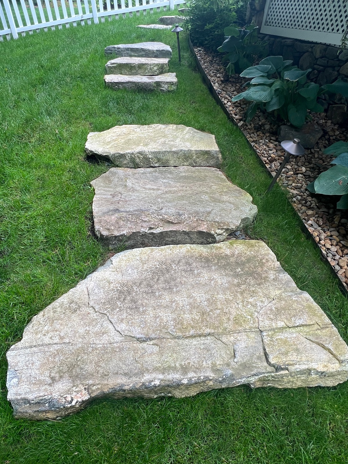 Stone stepping path leads through lush garden, bordered by white fence and small plants, with path lighting along landscaped edges.