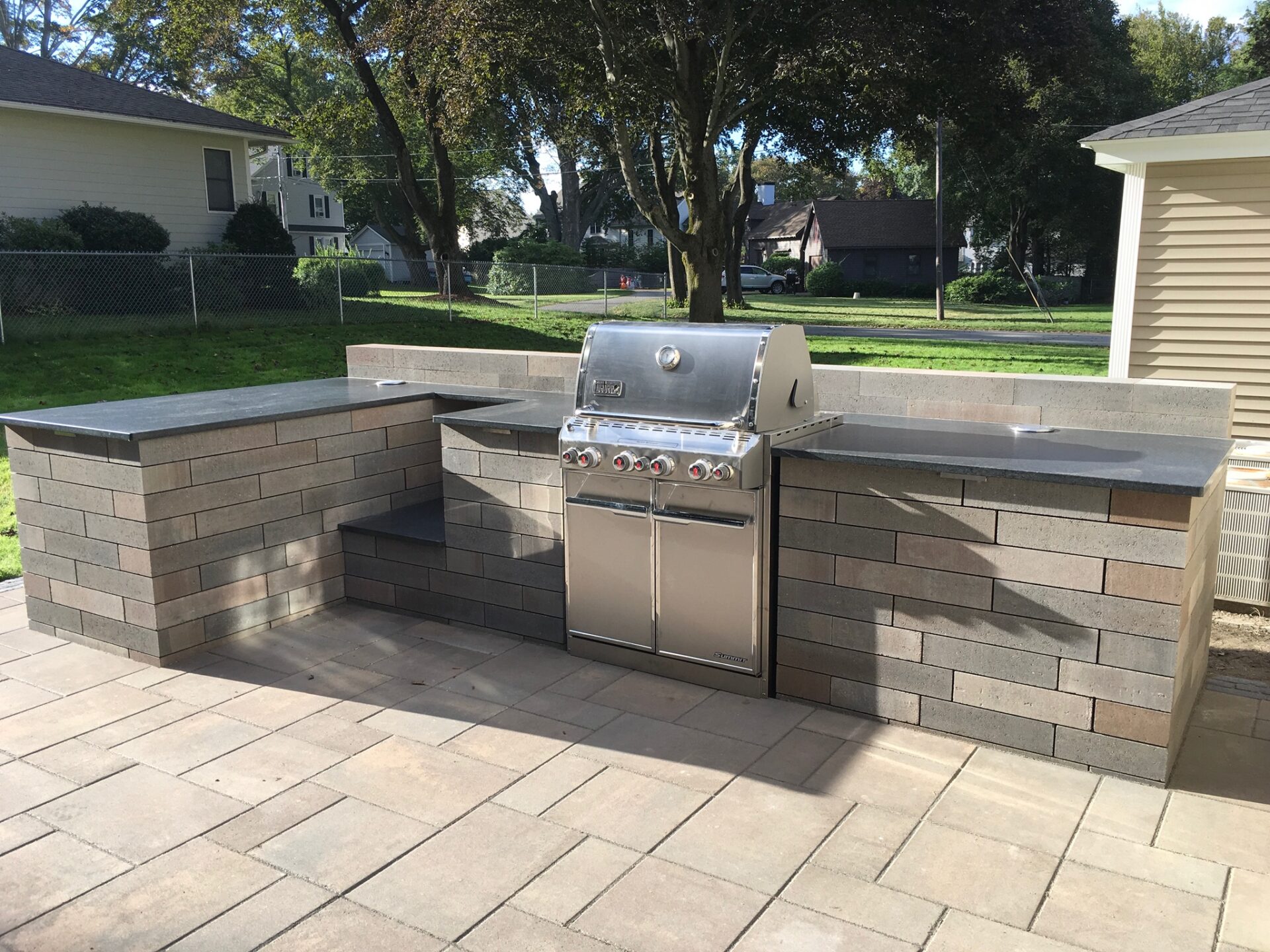 Outdoor kitchen with a stainless steel grill, surrounded by brick countertops on a spacious patio, set amidst a residential backyard.