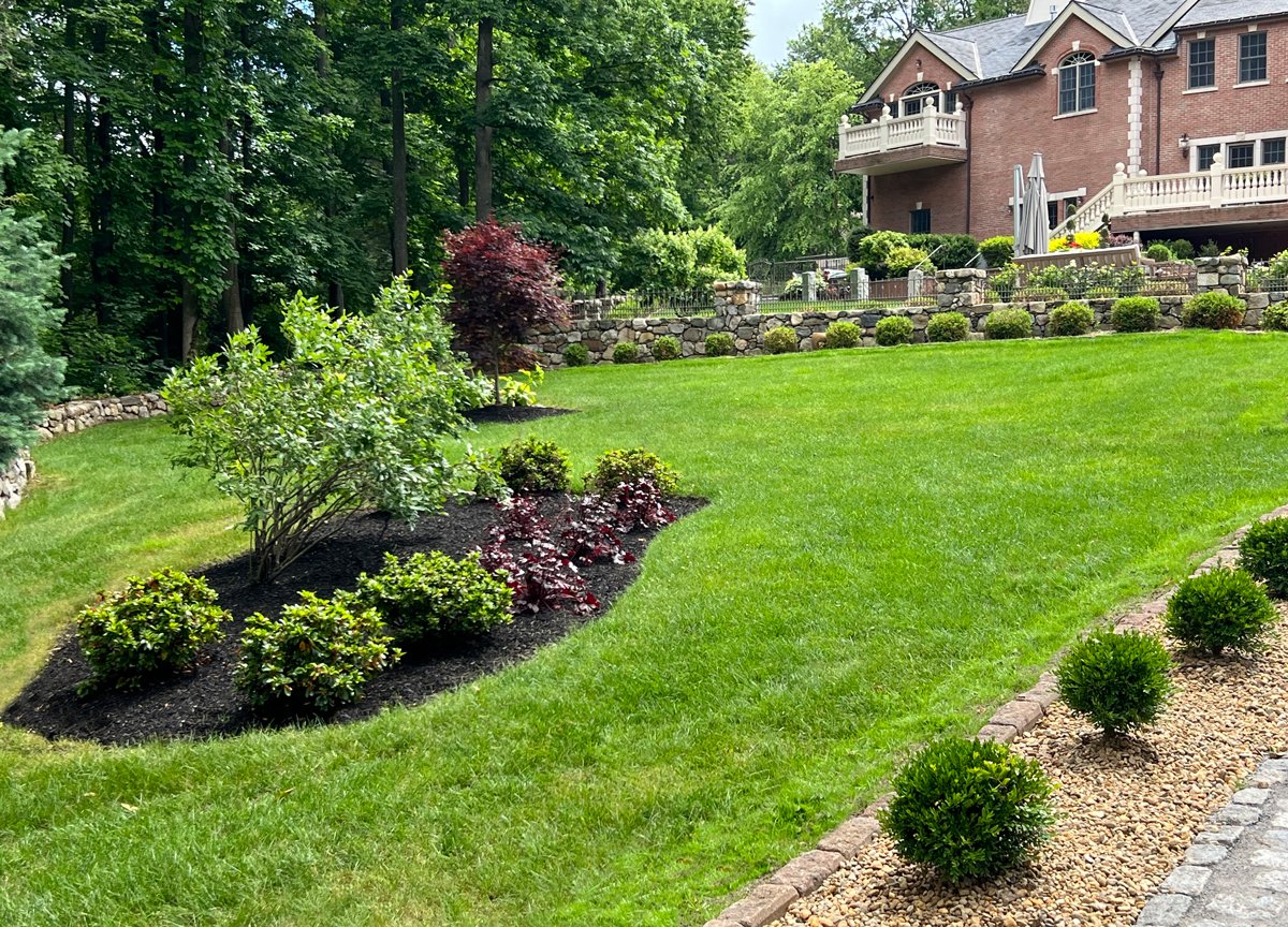Luxurious backyard with manicured lawn, shrubs, stone retaining walls, and a large brick house with balconies, surrounded by lush green trees.