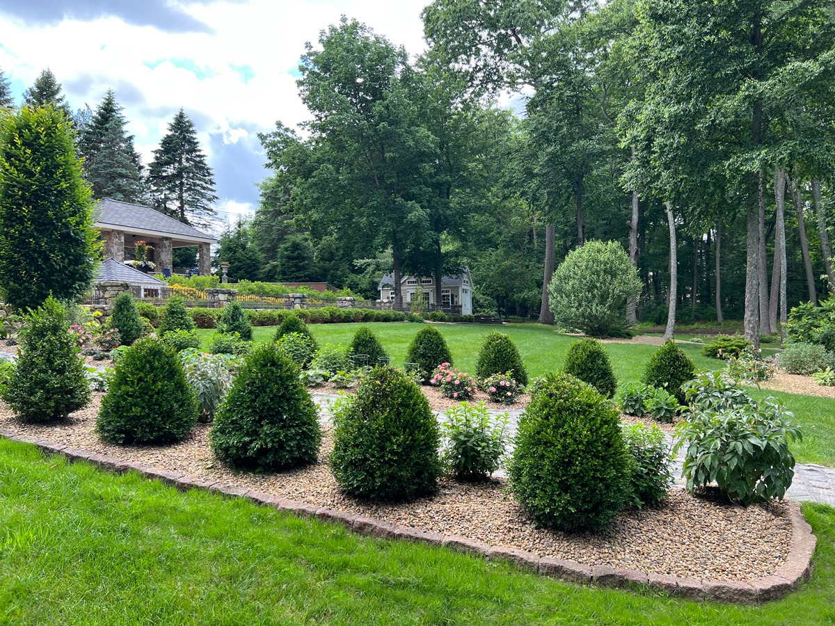 A well-manicured garden features neatly trimmed hedges, colorful flowers, and a stone pathway, surrounded by lush trees and a charming gazebo.