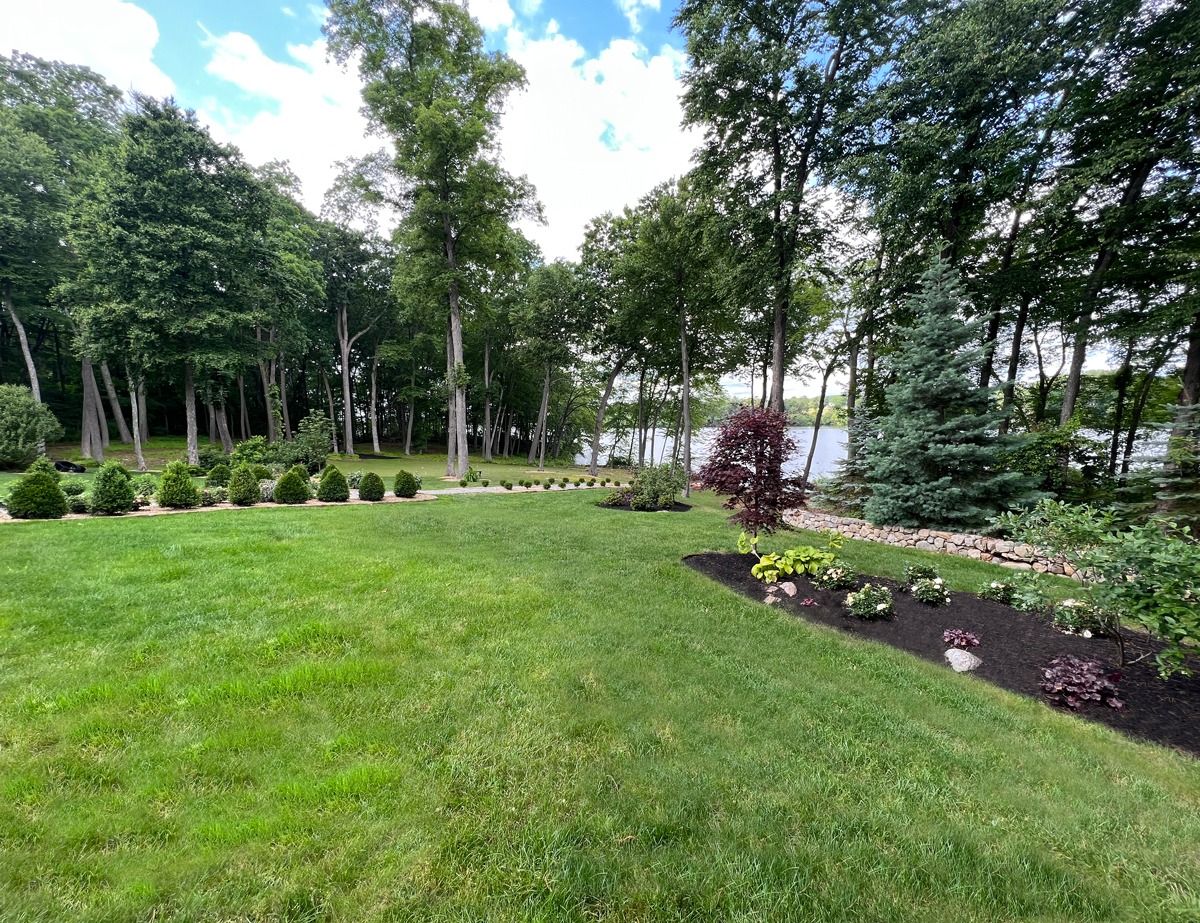 Lush green lawn with scattered flower beds, surrounded by tall trees. A serene lake is visible in the background under a partly cloudy sky.