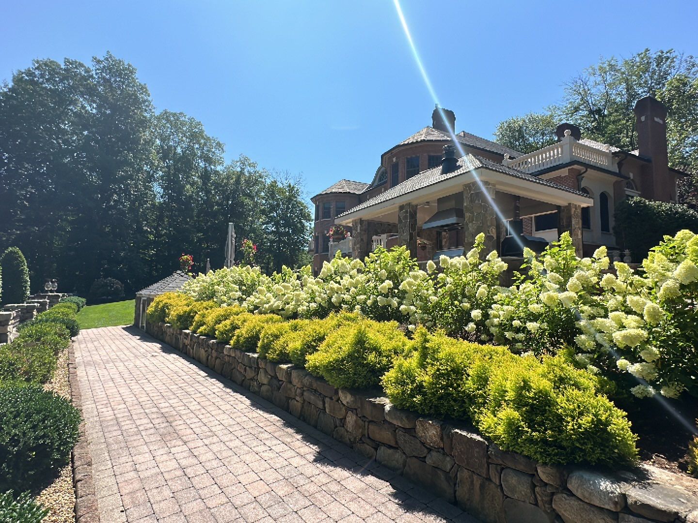 Stone path leads to a grand house with lush gardens and bright hydrangeas under a clear blue sky, surrounded by dense trees.