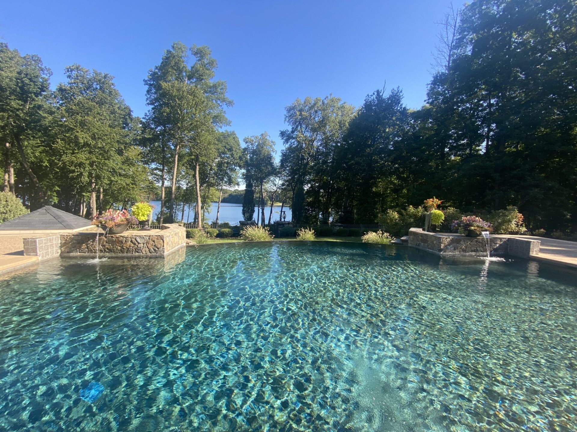 A serene pool with clear water, surrounded by lush trees, overlooks a tranquil lake under a bright, clear blue sky.
