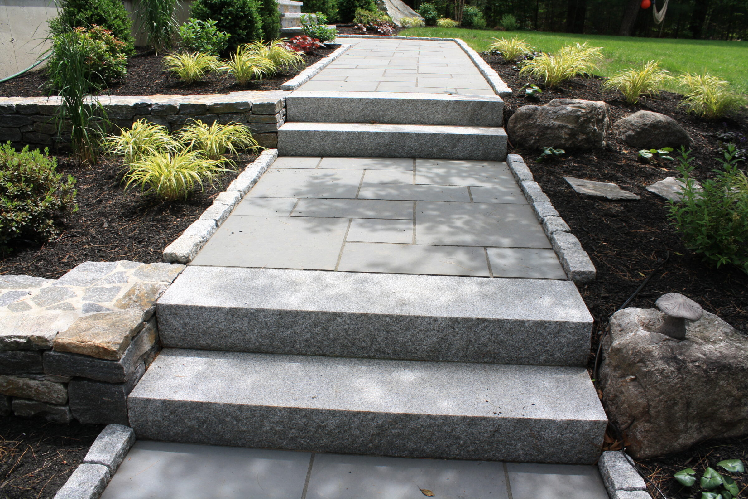 A stone pathway with steps, surrounded by neatly landscaped greenery and rocks, leads up to an unseen destination.