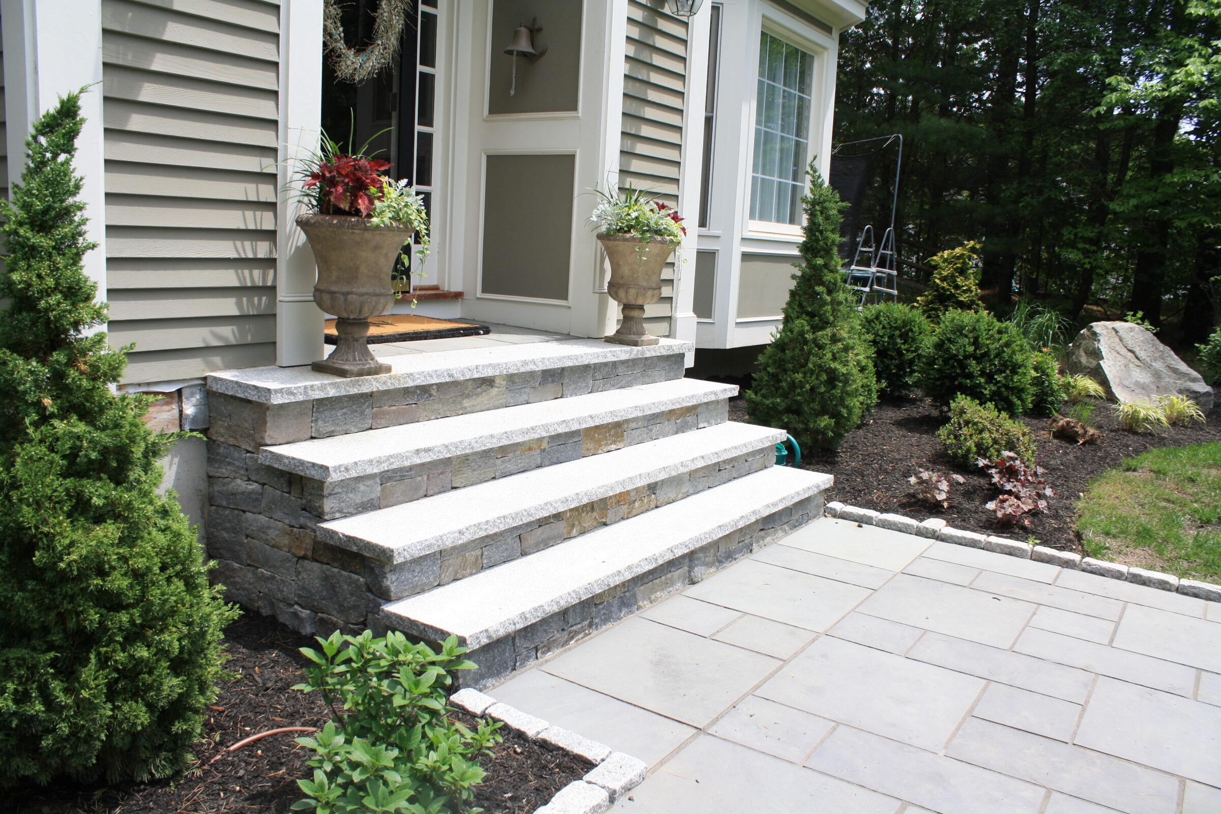 Stone steps lead to a house entrance with potted plants, surrounded by neatly landscaped greenery and a paved patio area.