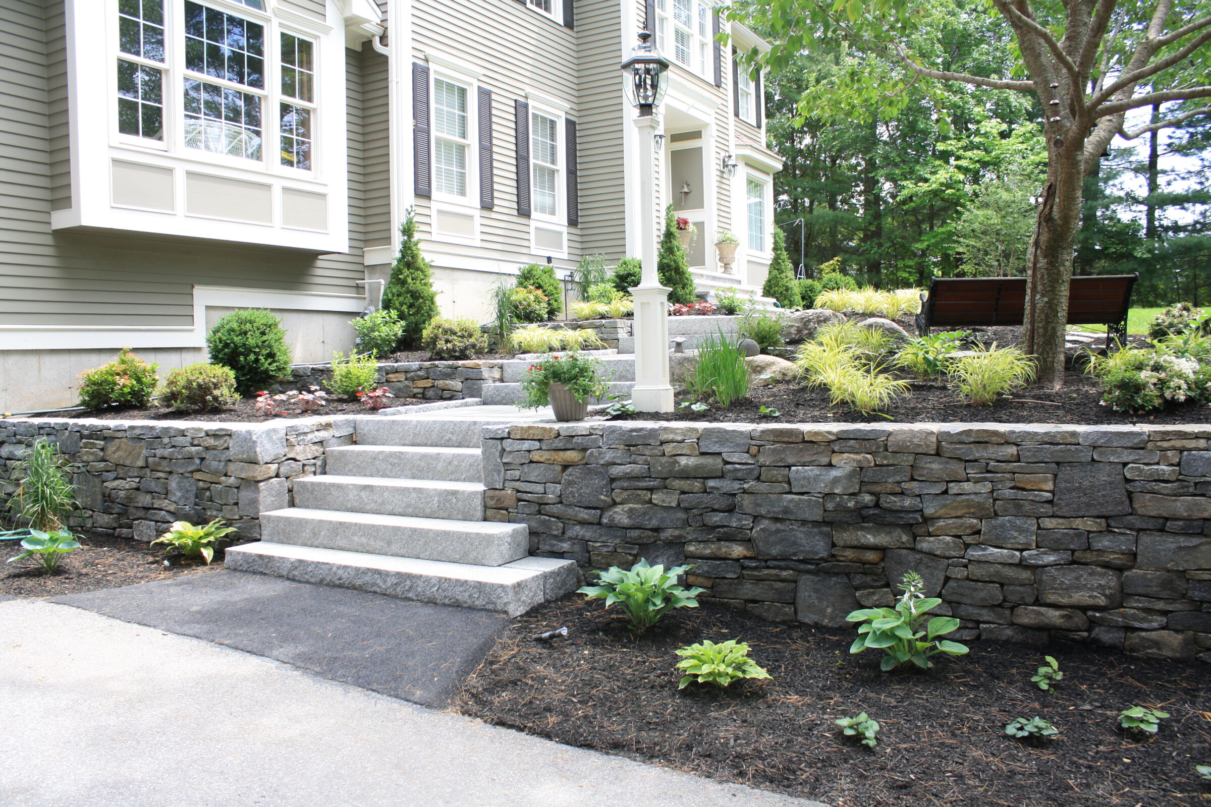 A well-maintained garden with stone walls, steps, and lush greenery surrounds the entrance of a two-story residential building under trees.