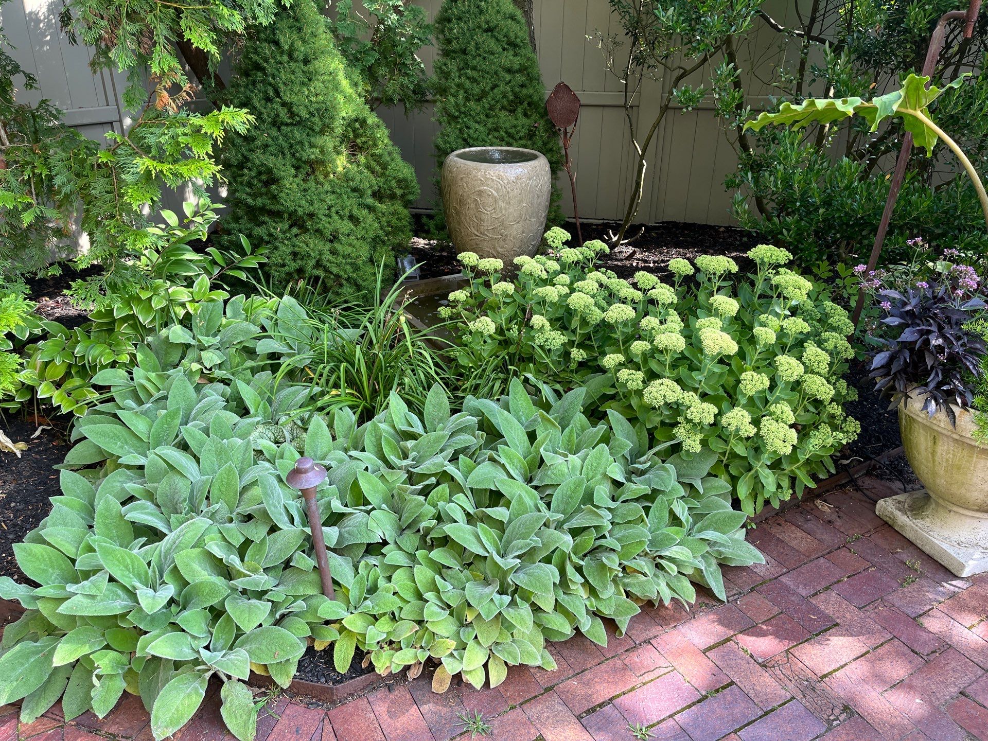 A serene garden scene with diverse green plants, a large ceramic pot, and a brick pathway in a peaceful courtyard setting.