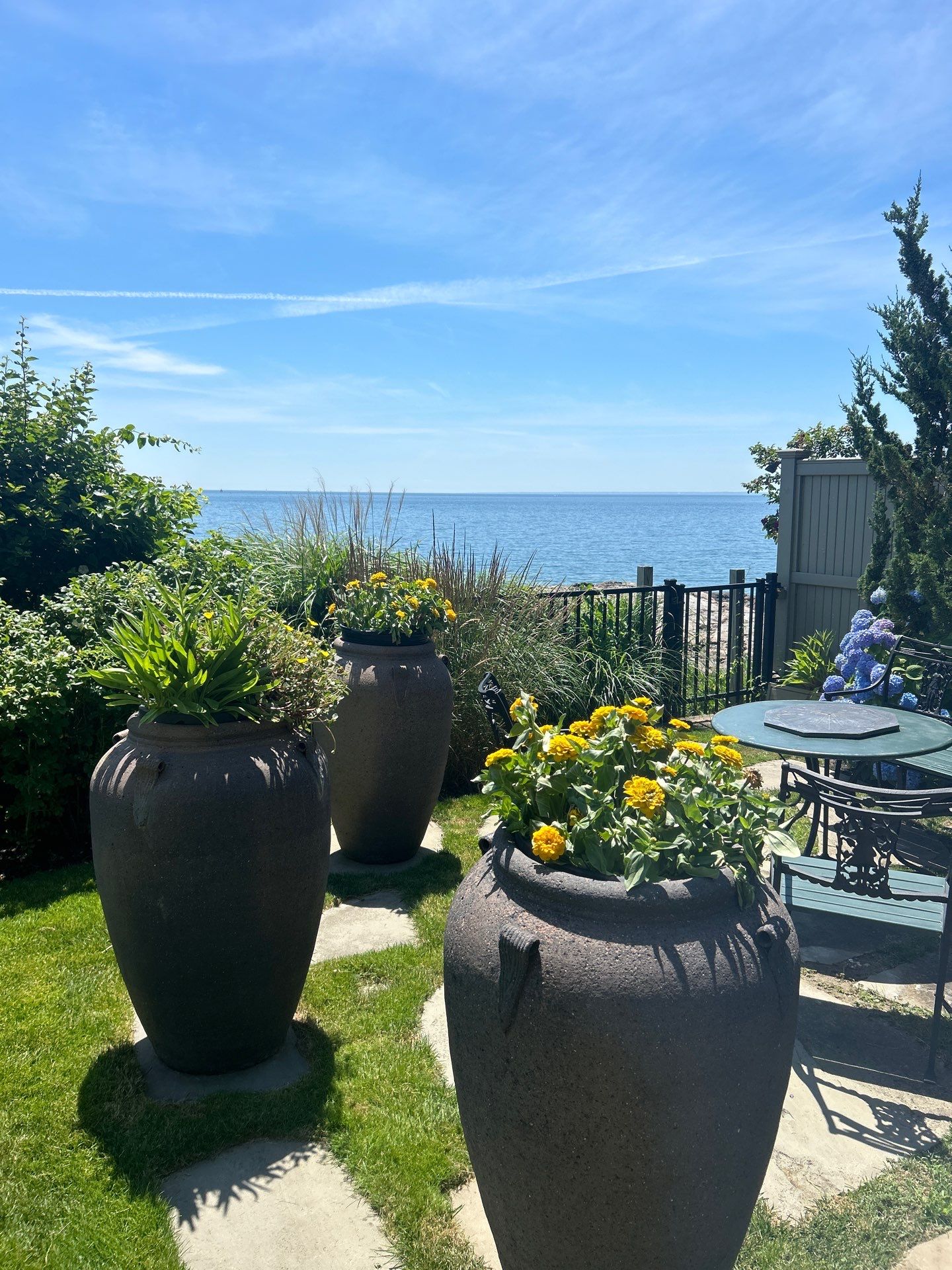 Large ceramic pots with flowers sit on a sunny garden patio, overlooking a serene ocean view under a clear blue sky.