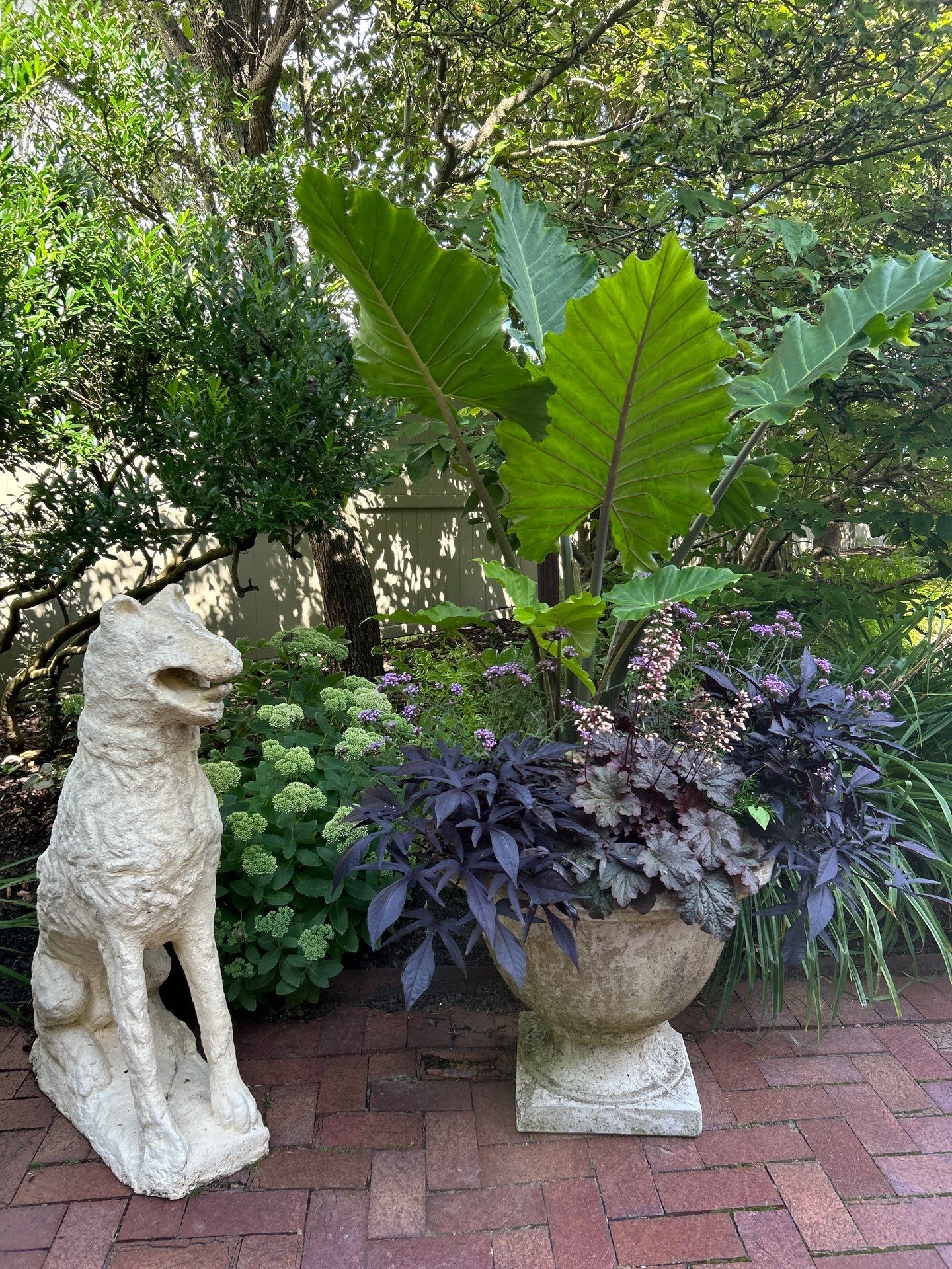 A garden scene with a dog statue and a large potted plant surrounded by greenery on a brick pathway.