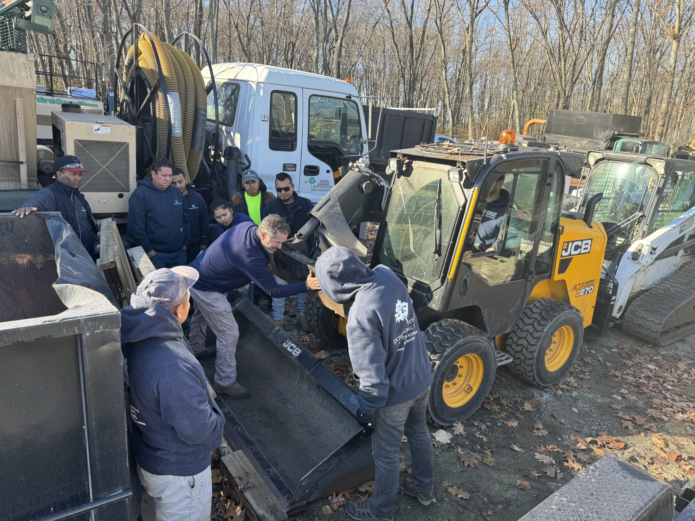 A group of people work with heavy machinery in an outdoor area surrounded by bare trees, observing and assisting with equipment handling.