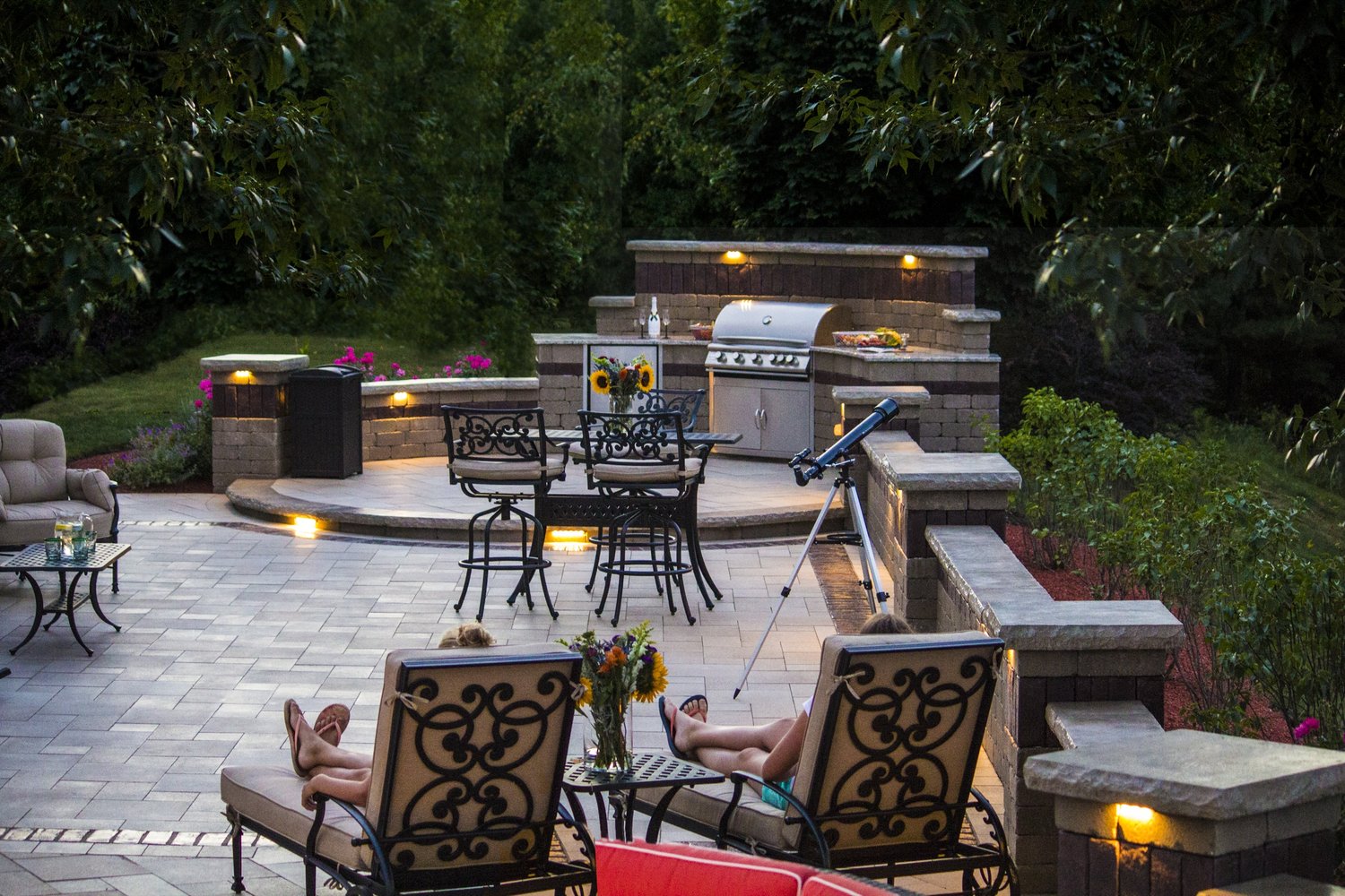 A cozy patio with elegant seating, two people relaxing, a telescope, a barbecue grill, and ambient lighting surrounded by lush greenery.