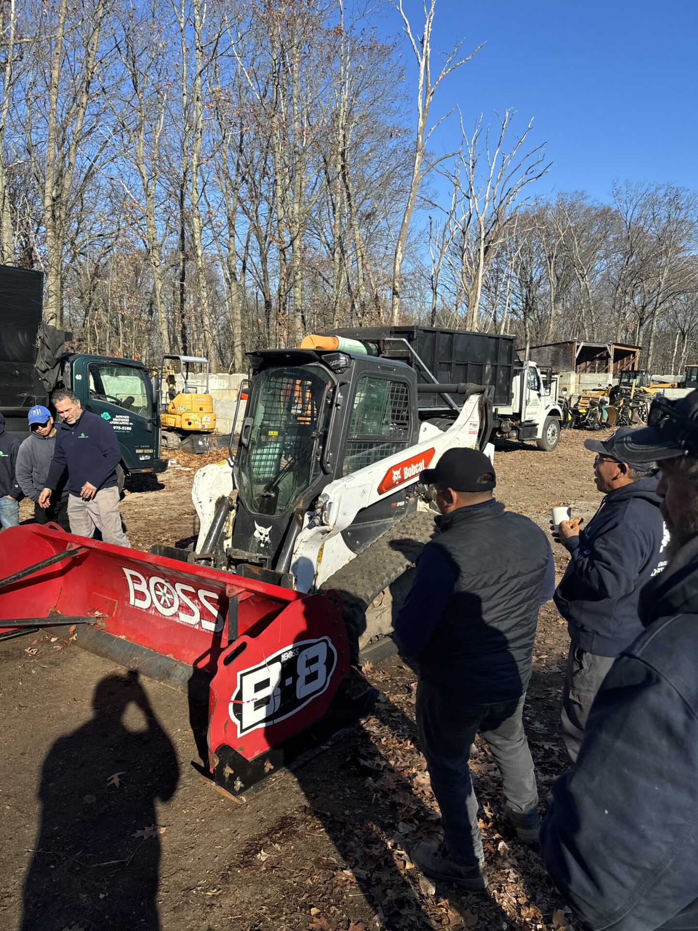 Four people stand near construction vehicles and a red snowplow attachment in a wooded area. It's a clear, sunny day.