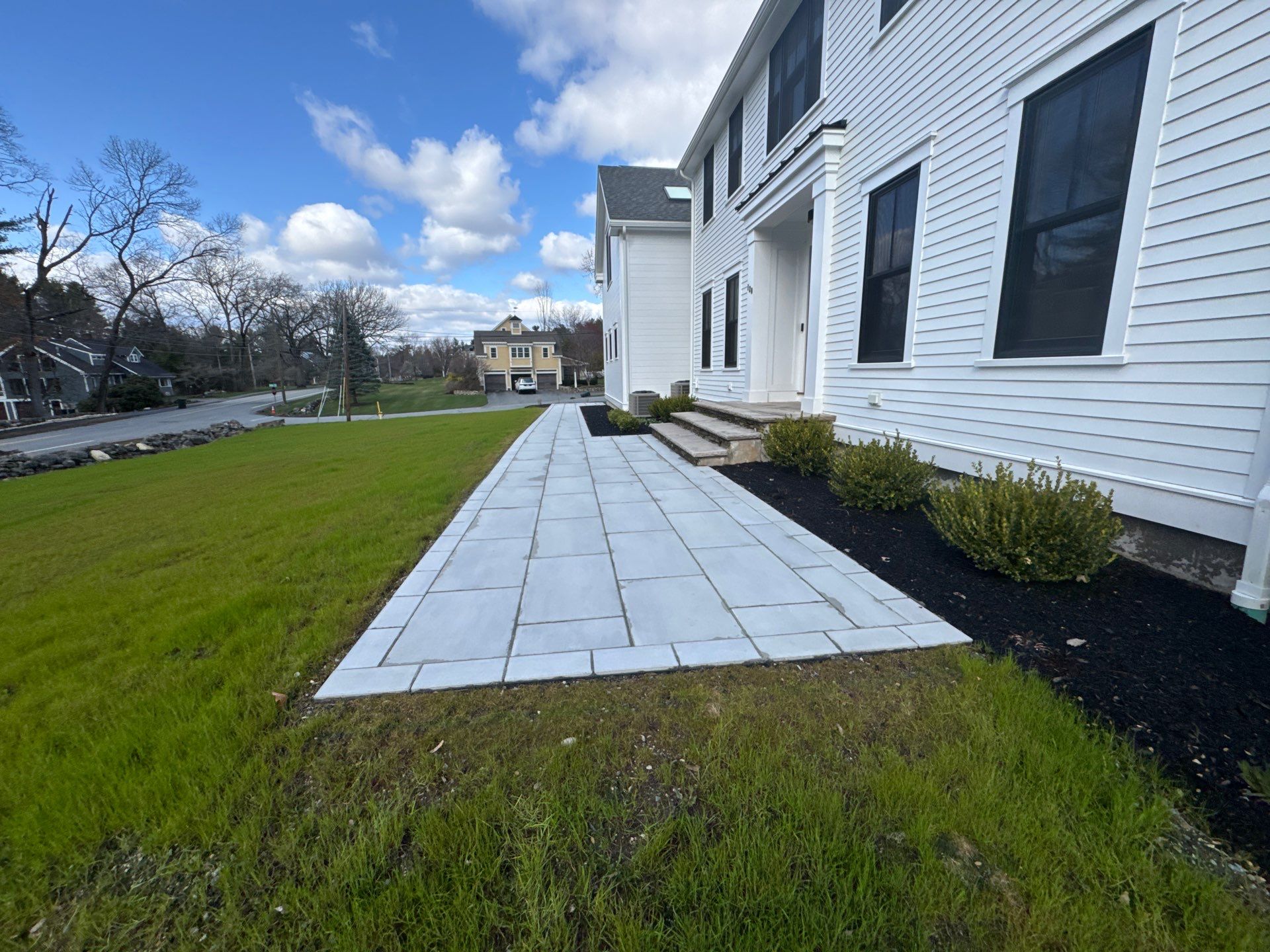 White house with paved pathway, green lawn, and bushes. Background includes trees, a road, and another house in suburban setting under a blue sky.