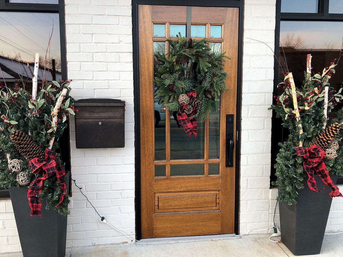 A wooden door adorned with a festive wreath, flanked by decorative arrangements, creates a welcoming holiday entrance on a white brick wall.