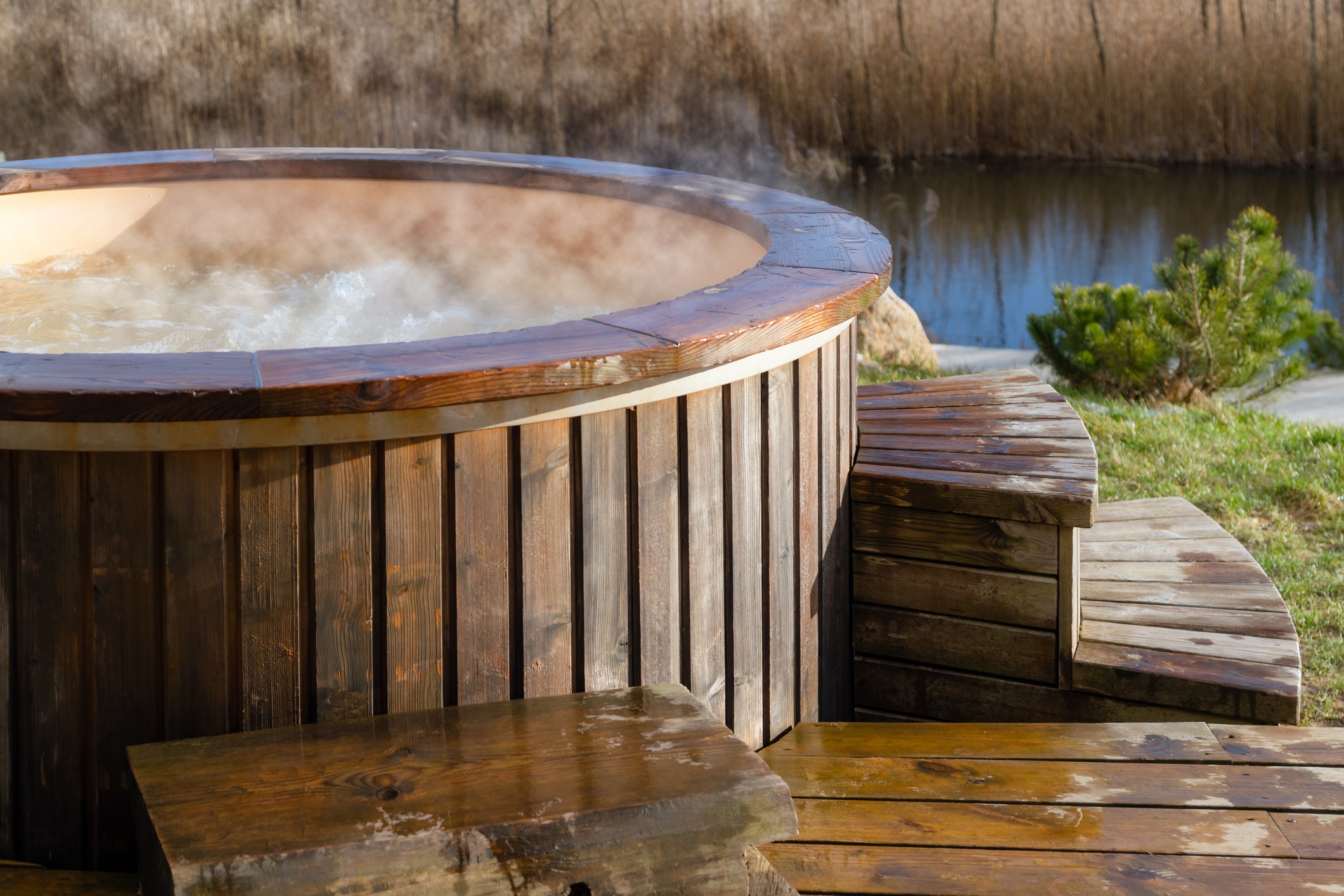 A wooden hot tub with steaming water, surrounded by wooden steps, sits beside a calm river and grassy area.