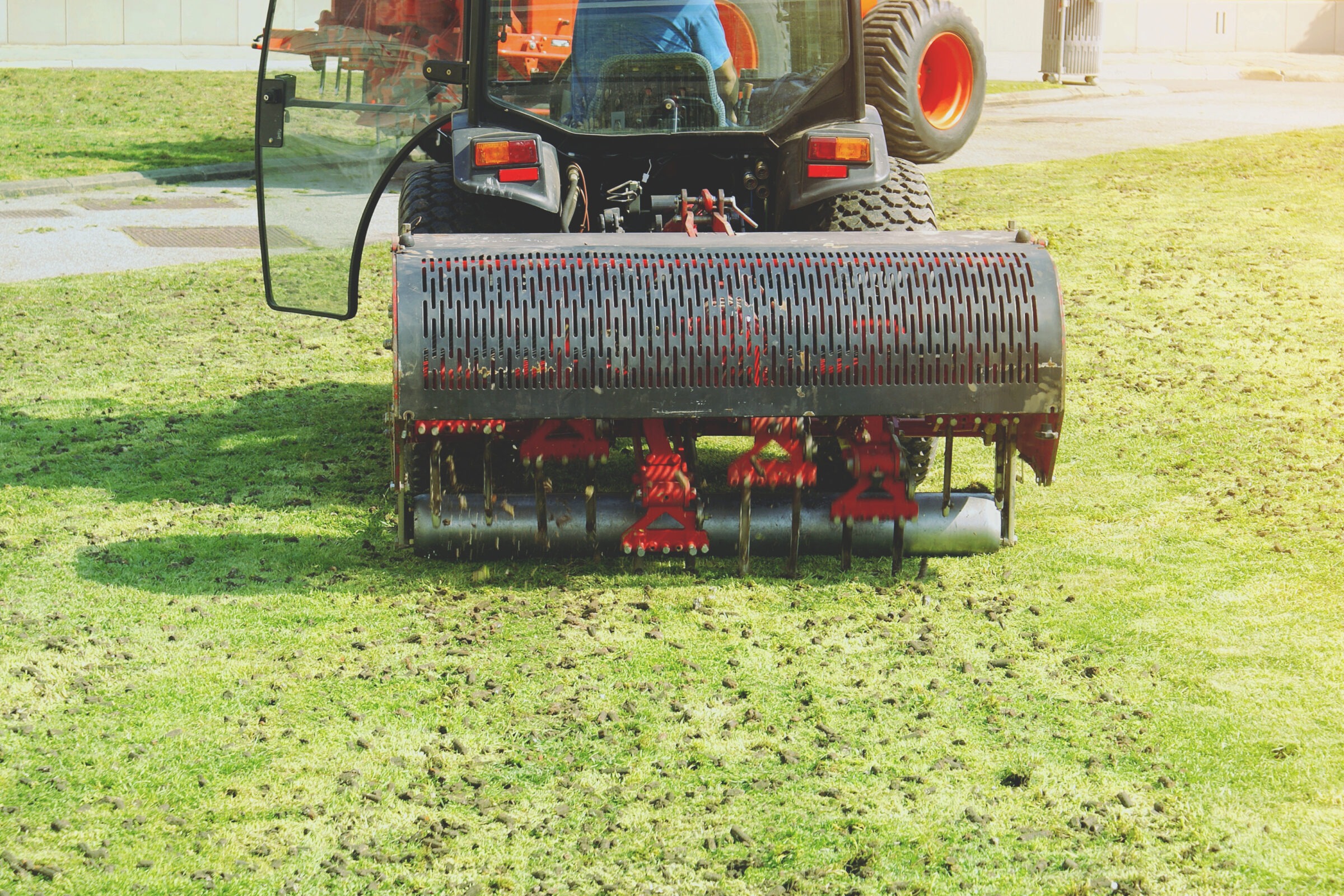 A tractor aerates a grassy field with a spiked roller attachment, operated by a person in a blue shirt, in a sunny setting.