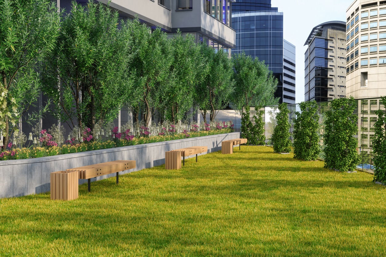 Rooftop garden with wooden benches, lush greenery, and colorful flowers, surrounded by modern skyscrapers in an urban setting.