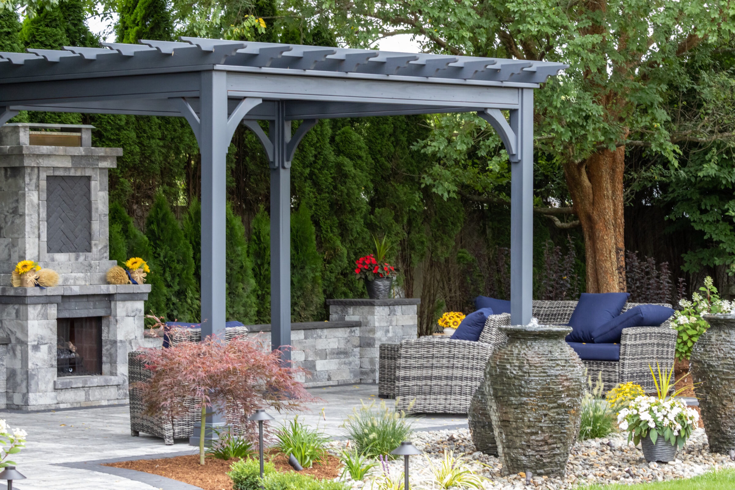 A garden patio features a gray pergola, stone fireplace, wicker furniture with blue cushions, potted plants, and lush greenery in the background.