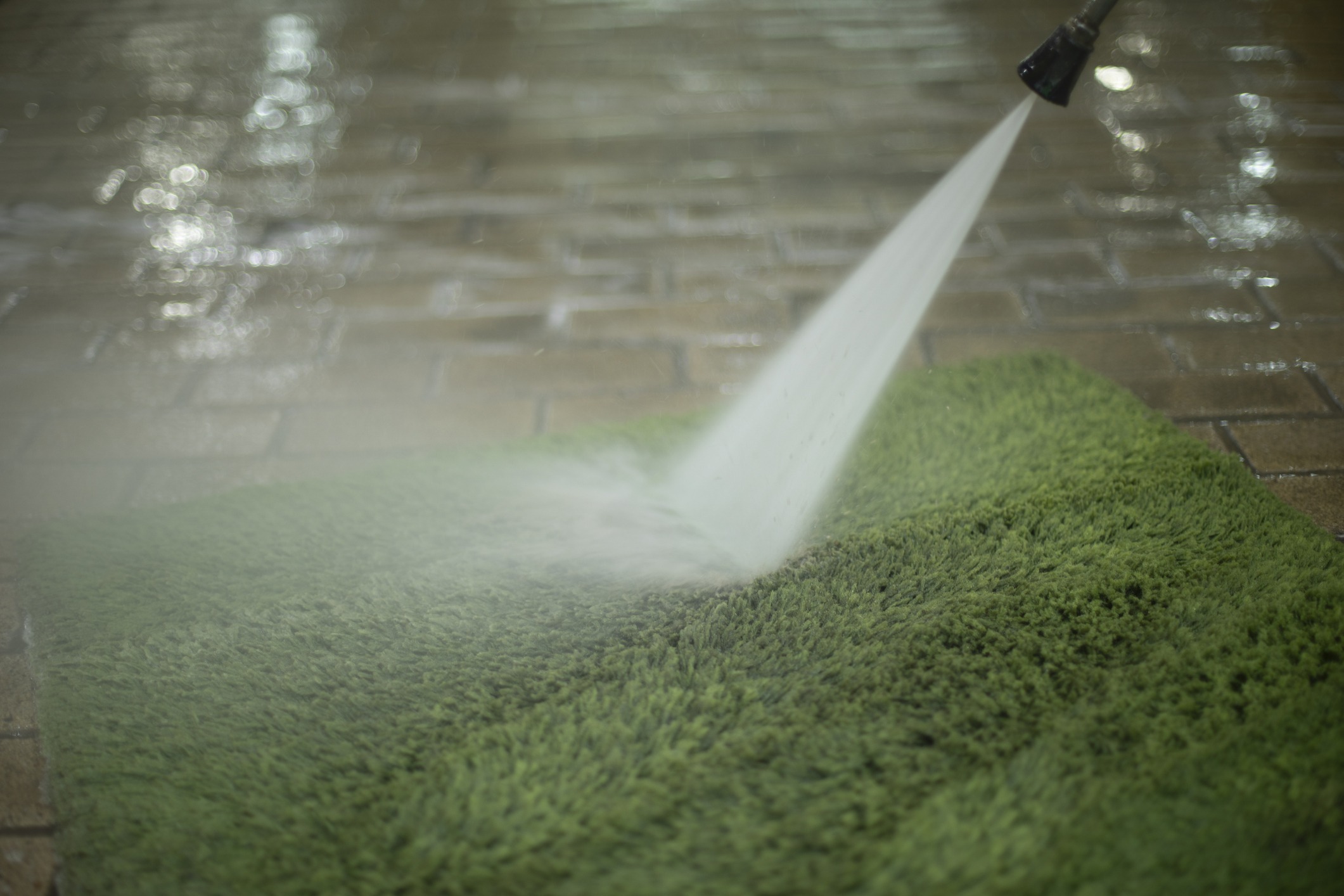 A high-pressure water spray cleans a green outdoor mat on a wet stone surface, creating a misty, fresh, and tidy environment.