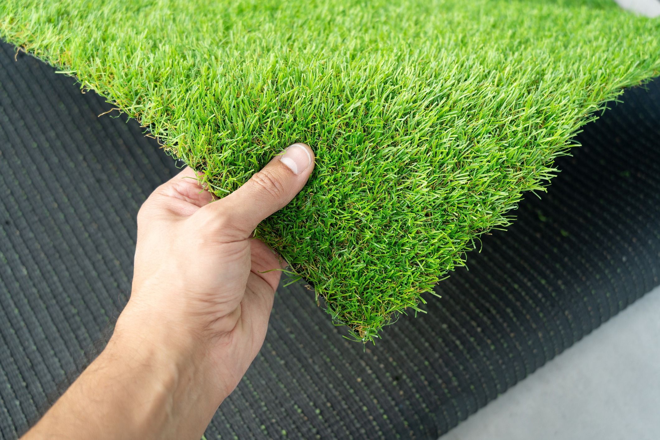 A person holds a piece of artificial grass, showcasing its vibrant green texture against a dark, ribbed background.
