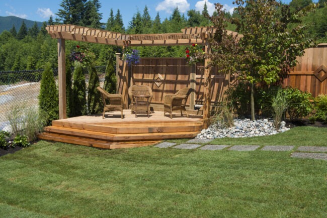 A small wooden patio and pergola in a lush green backyard.