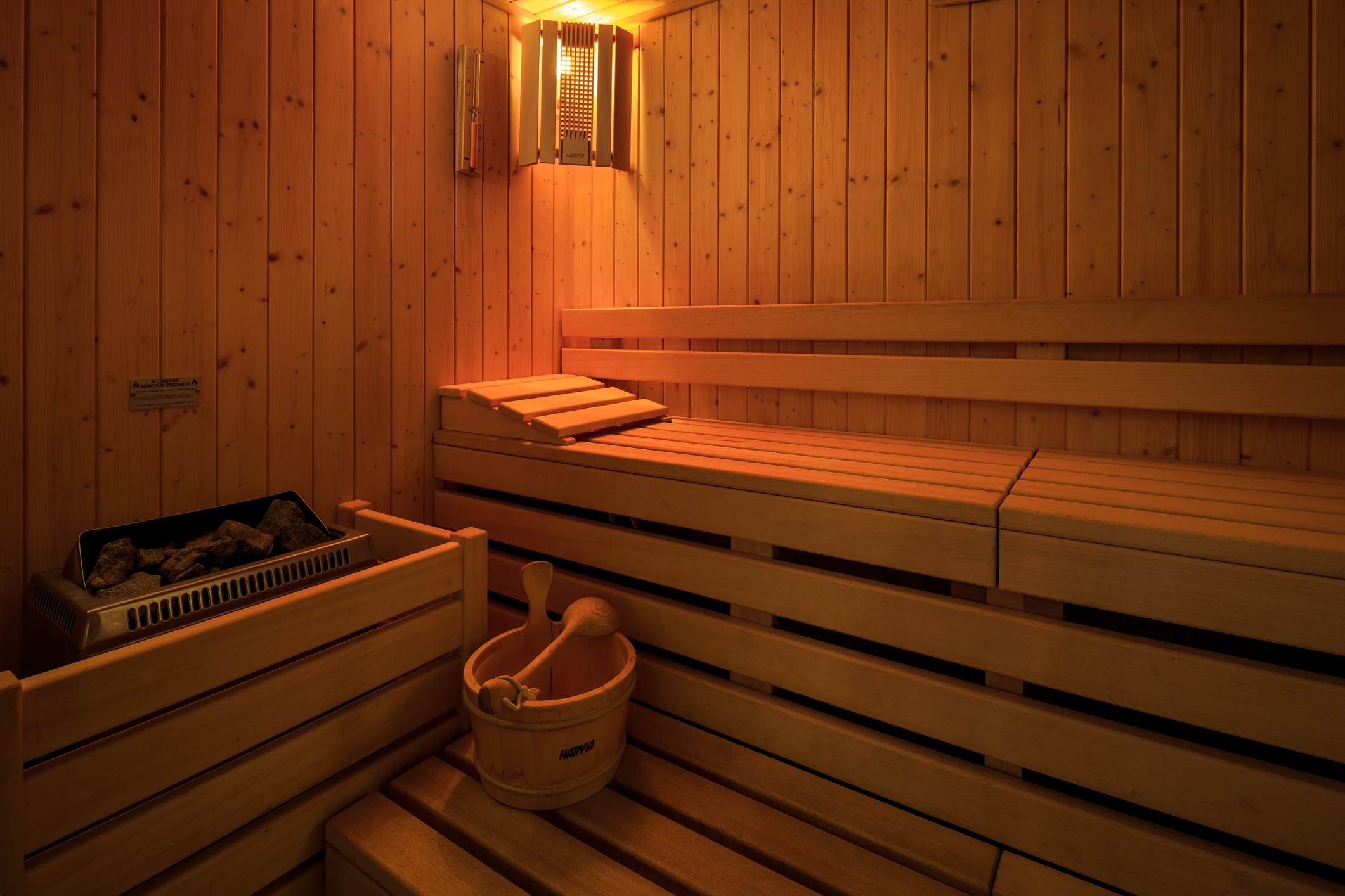 Wooden sauna interior with benches, rocks in a heater, and a wooden bucket and ladle. Warm ambient lighting enhances a tranquil atmosphere.