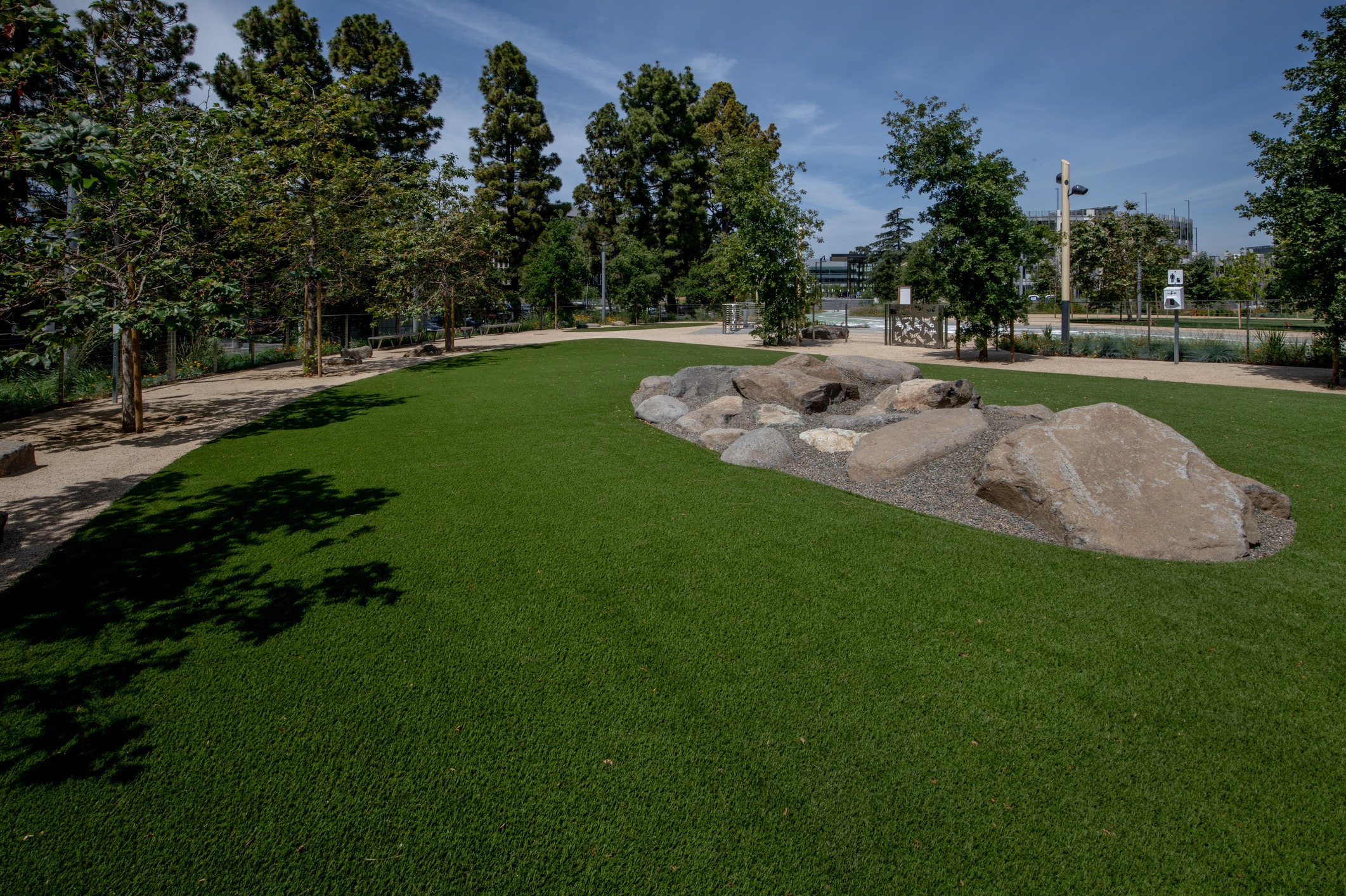 Lush park scene featuring green grass, large rocks, and surrounding trees under a clear blue sky. Pathway winds through the landscaped area.