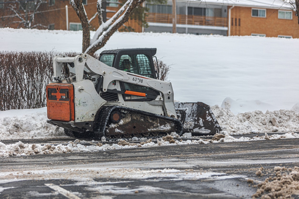 A person operates a snow plow in a snowy residential area, clearing roads. Brick buildings and trees are visible in the background.