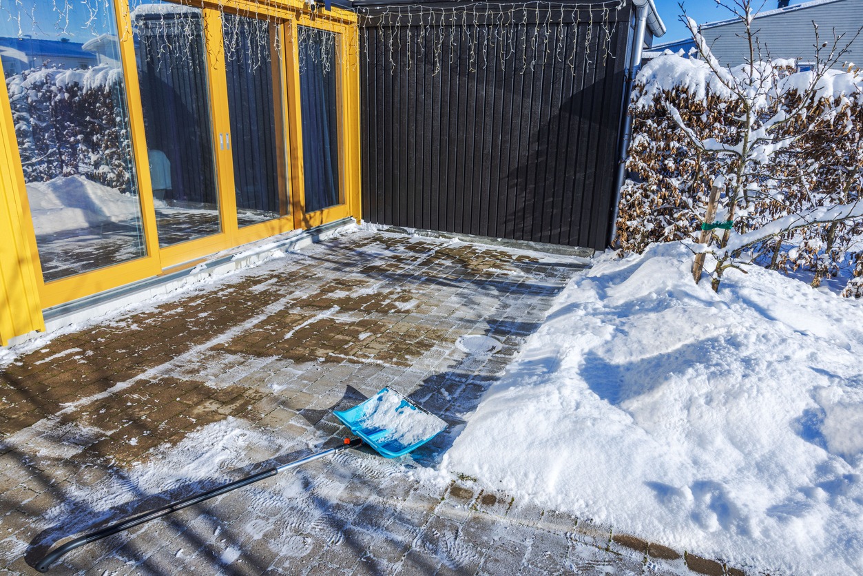 Bright patio with yellow sliding doors, partially covered in snow. Snow shovel left on cleared brick path. Snow-covered hedge and tree nearby.