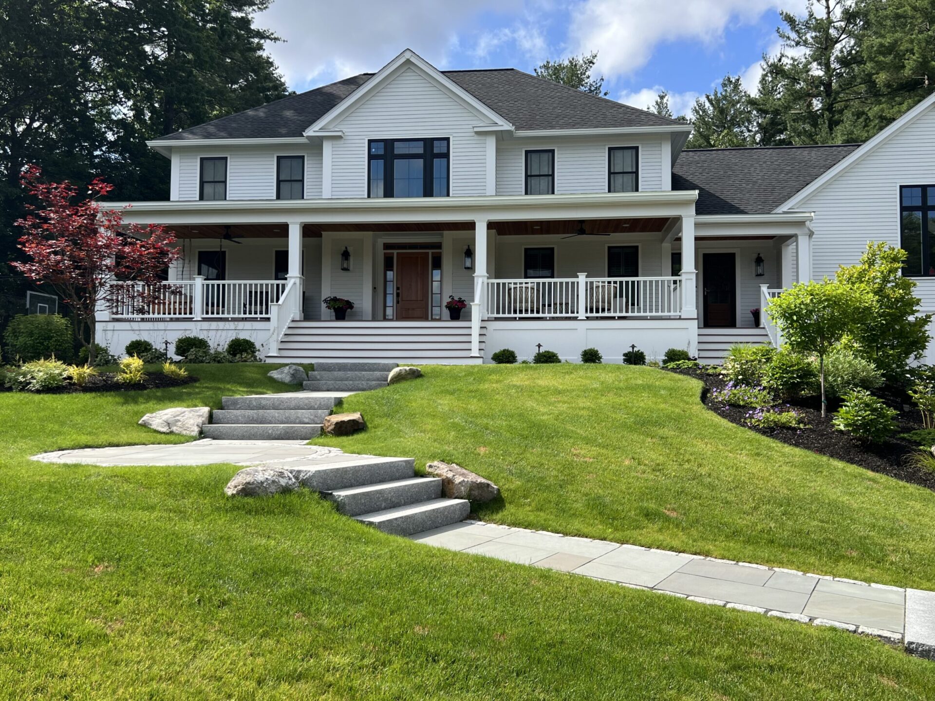 A well-maintained lawn with landscaping leads follows a stone paver path to a house featuring white railings, steps, and a porch, surrounded by lush greenery and trees.